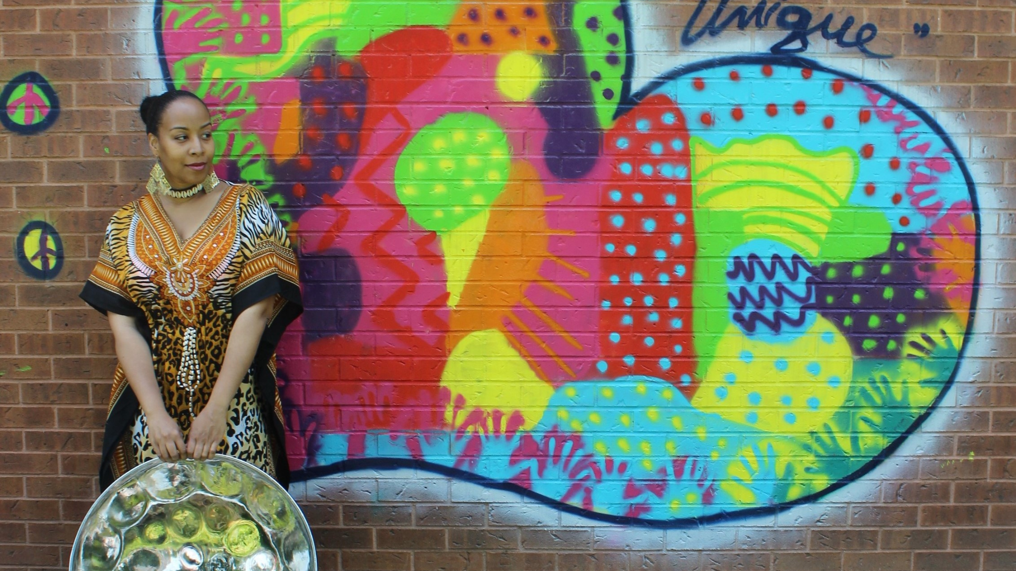 Black woman holding a steelpan in front of a colourful heart painted on a brick all