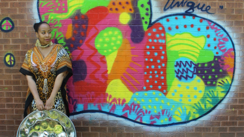 Black woman holding a steelpan in front of a colourful heart painted on a brick all