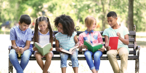 Kids sitting on a bench outside reading a book.