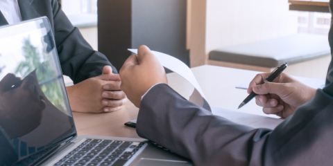 laptop on a table beside a person's hands holding a pen about to write in a notebook