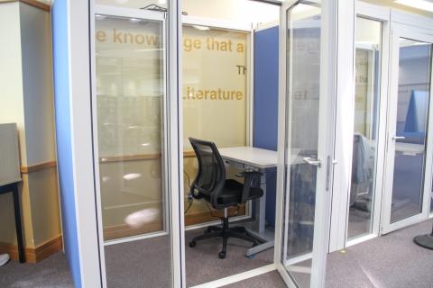 A picture of a work pod with glass windows and the door open. Inside is a desk and chair.