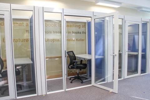 A picture of a work pod with glass windows and the door open. Inside is a desk and chair.