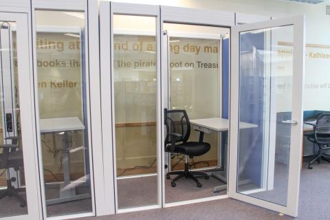 A picture of a work pod with glass windows and the door open. Inside is a desk and chair.