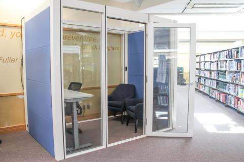 A picture of a work pod with glass windows and the door open. Inside is a desk and 3 chairs.