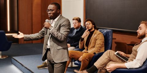 Man with arm extended, microphone in hand, three men behind him listening thoughtfully. 