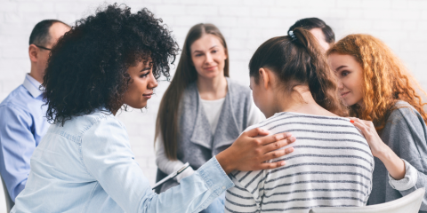 A group of people seated in a circle, one woman has a hand on another's shoulder.