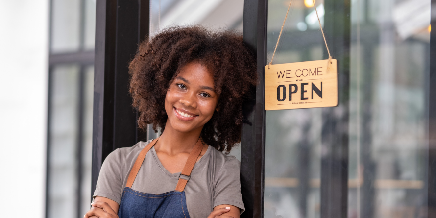 Smiling woman beside a "welcome, open" sign on a building. 