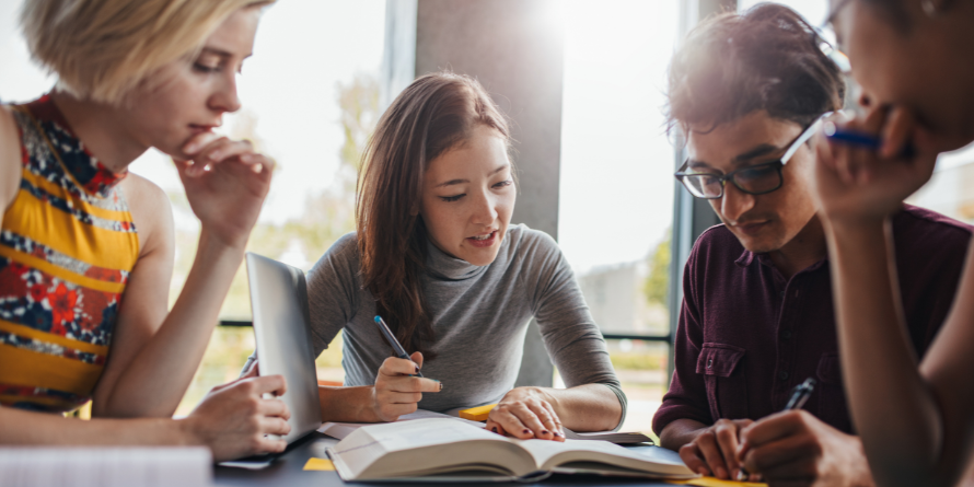 Four people gathered around a table, speaking, with papers and laptops out. 